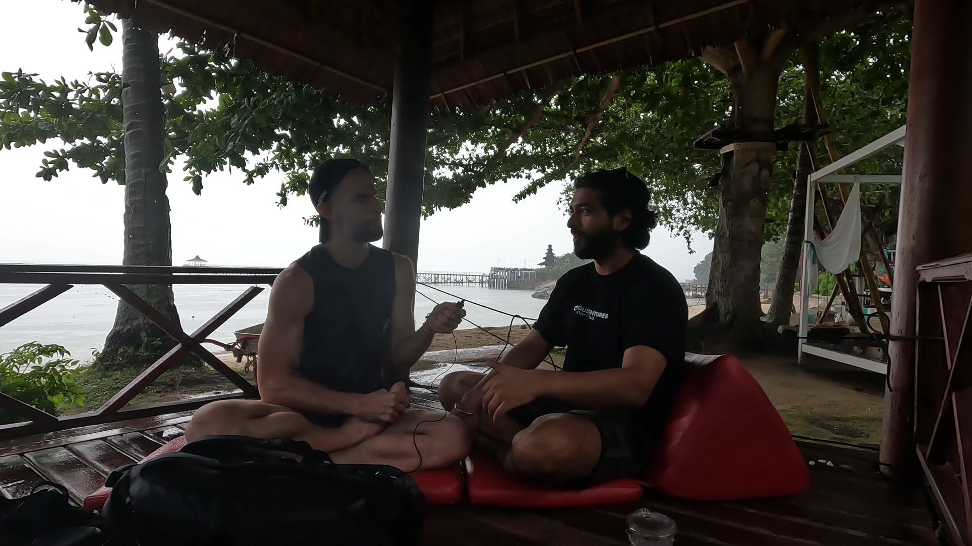 Ashish and Konrad sitting under an open-air hut by the sea at Network School, mid-conversation