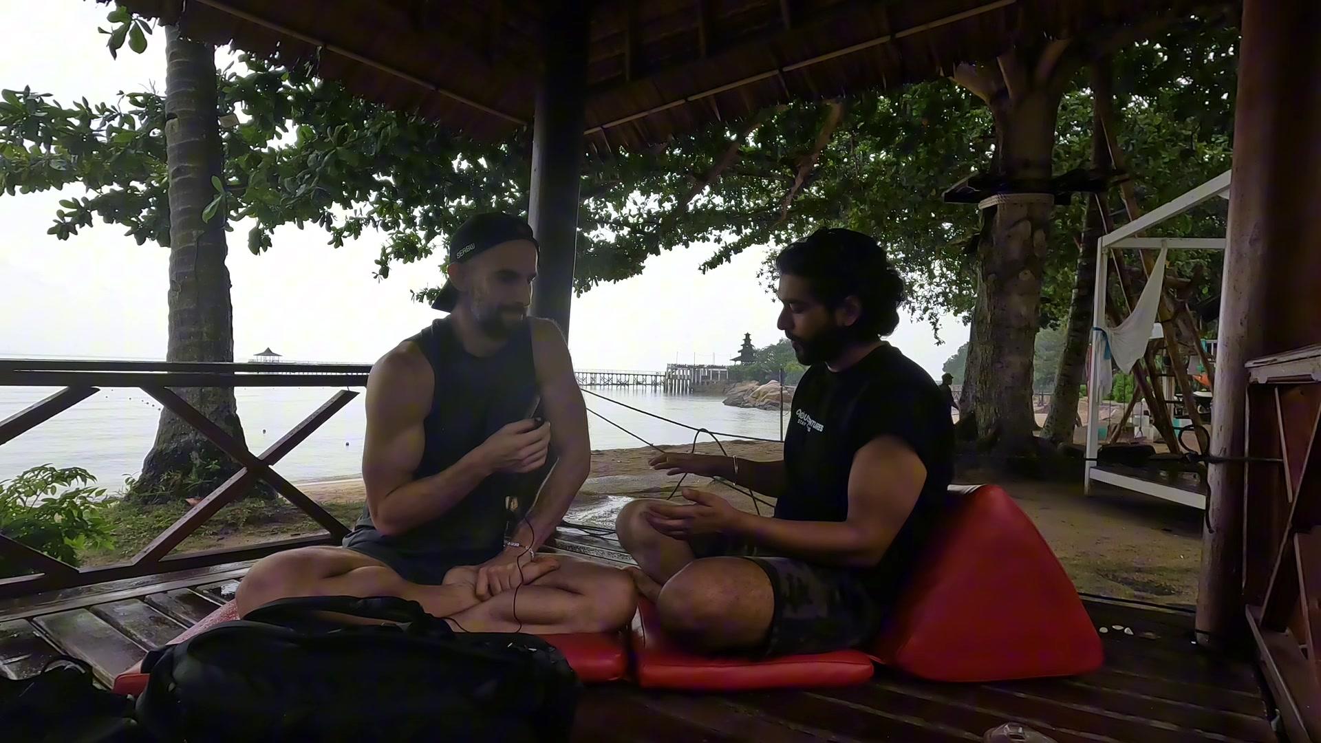 Ashish and Konrad seated cross-legged under the hut with the sea and a pier behind them