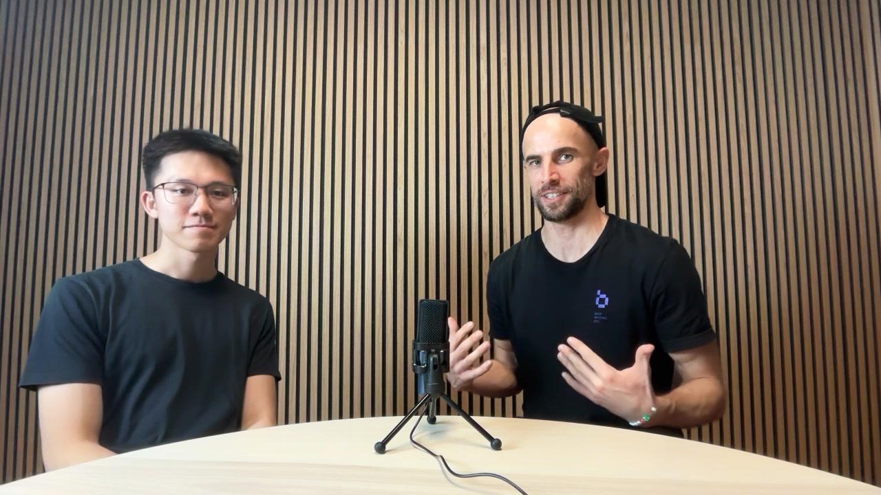 Two people sitting across a table with a microphone at Network School, at the start of a filmed conversation