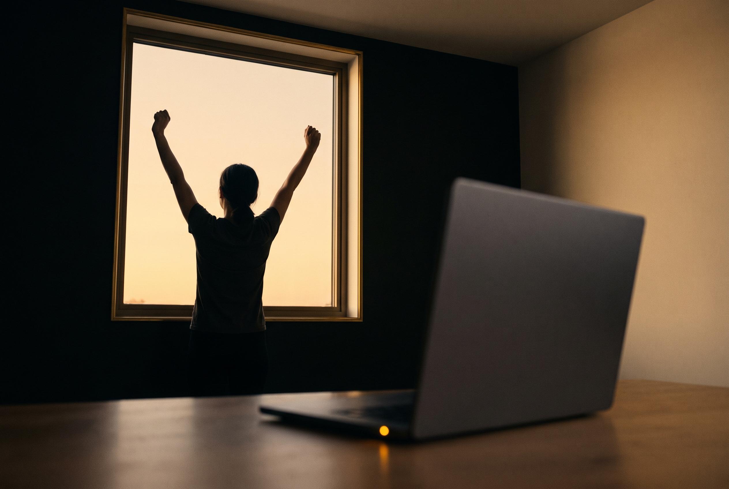 A person raising both arms at a window at dusk, away from a laptop resting on the desk