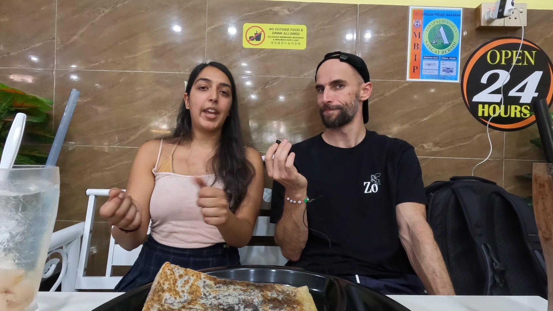 Velveteen (Miriam Reza) talking with her hands mid-gesture, and Konrad, during the podcast at a table in Forest City